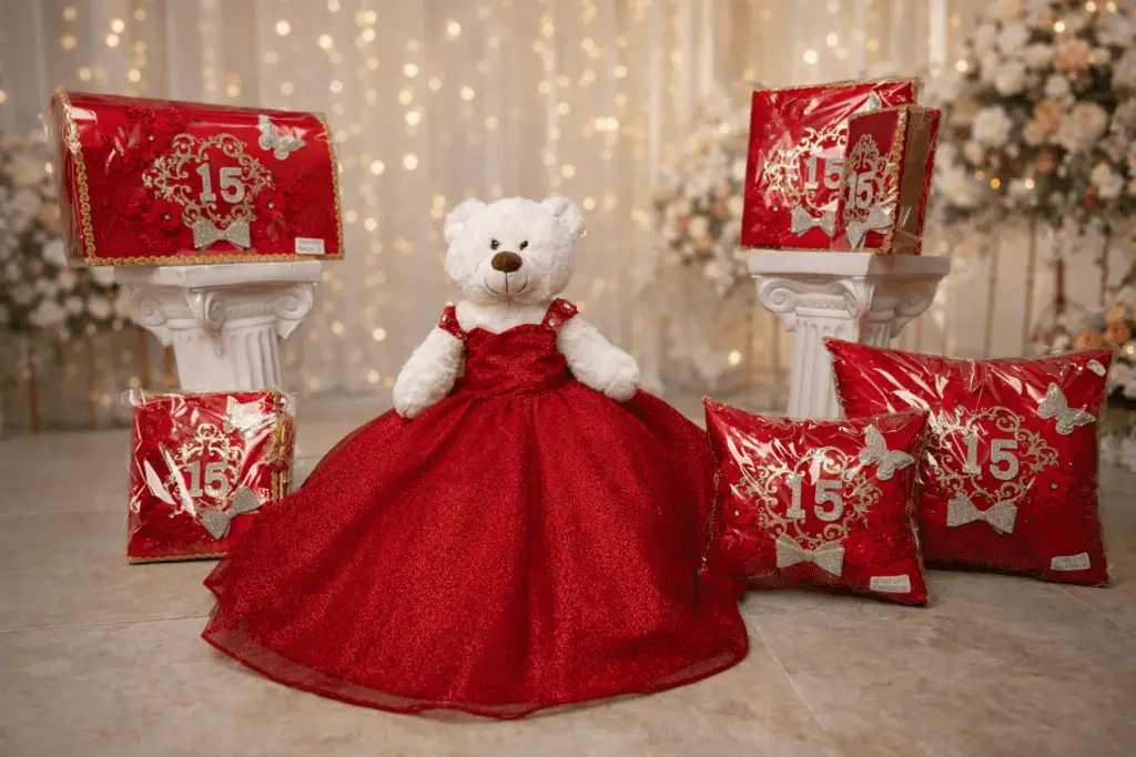 Oso de peluche con vestido rojo rodeado de cojines y cajas decorativas con número 15 para celebración de quinceañera.