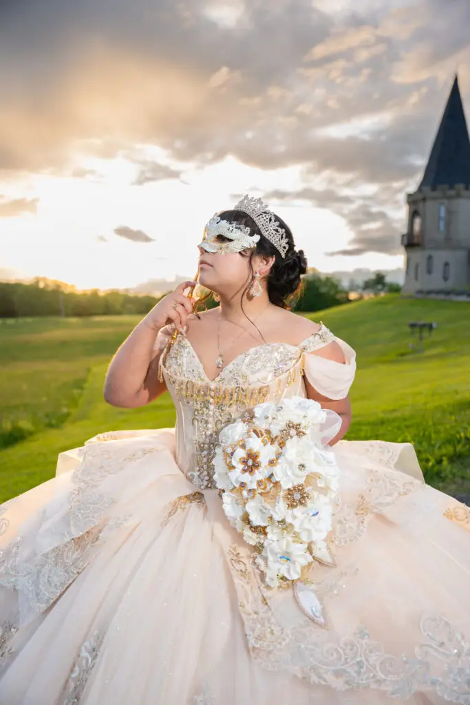 Quinceañera con vestido color champán y máscara decorativa, sosteniendo ramo blanco al atardecer en jardín.