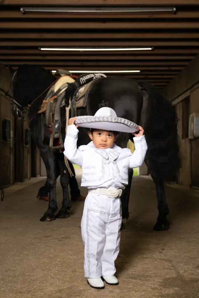 Niño con traje blanco estilo charro levantando sombrero, de pie frente a caballo negro en establo.