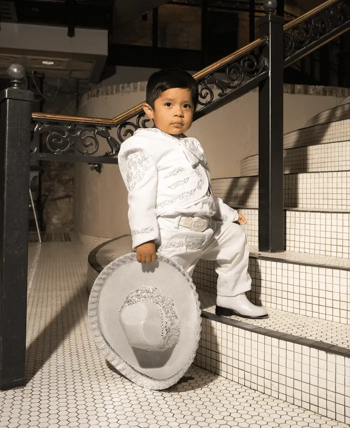 Niño con traje formal blanco estilo charro y sombrero, posando en escaleras con barandal decorativo.