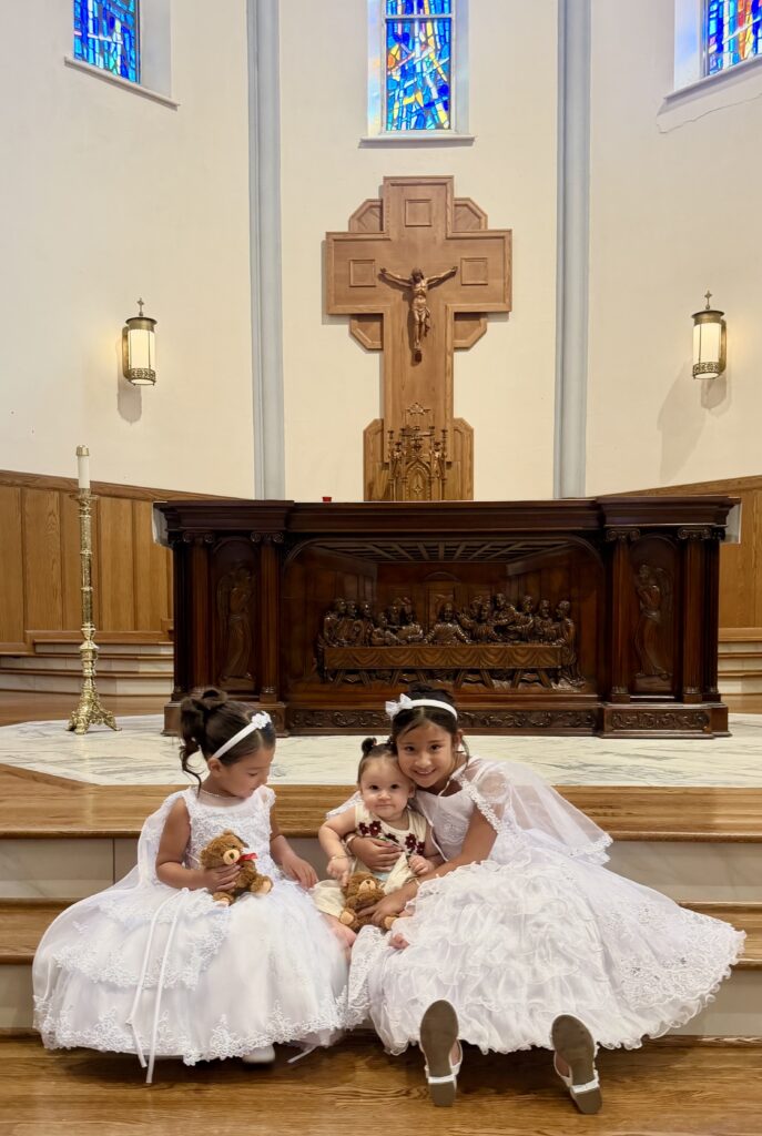 Tres niñas vestidas de blanco posando juntas frente al altar de una iglesia, luciendo vestidos con encaje y accesorios a juego.