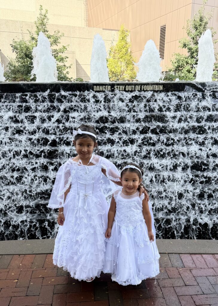 Dos niñas pequeñas sonriendo con vestidos blancos de presentación y bautizo con detalles de encaje y diademas frente a una fuente de agua.
