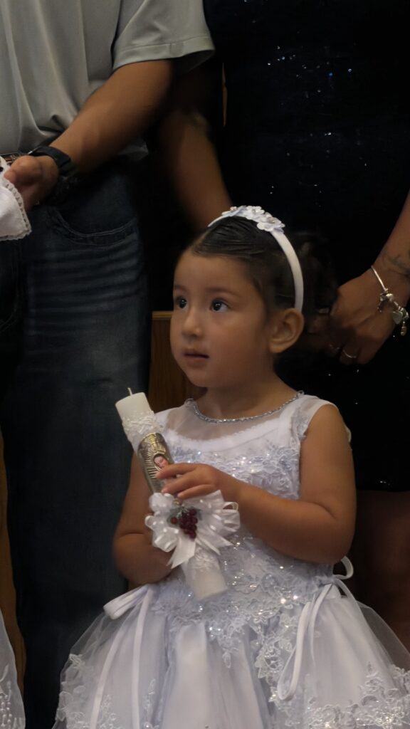 Niña pequeña en la iglesia luciendo vestido blanco con pedrería y diadema de flores mientras sostiene una vela de bautizo decorada.